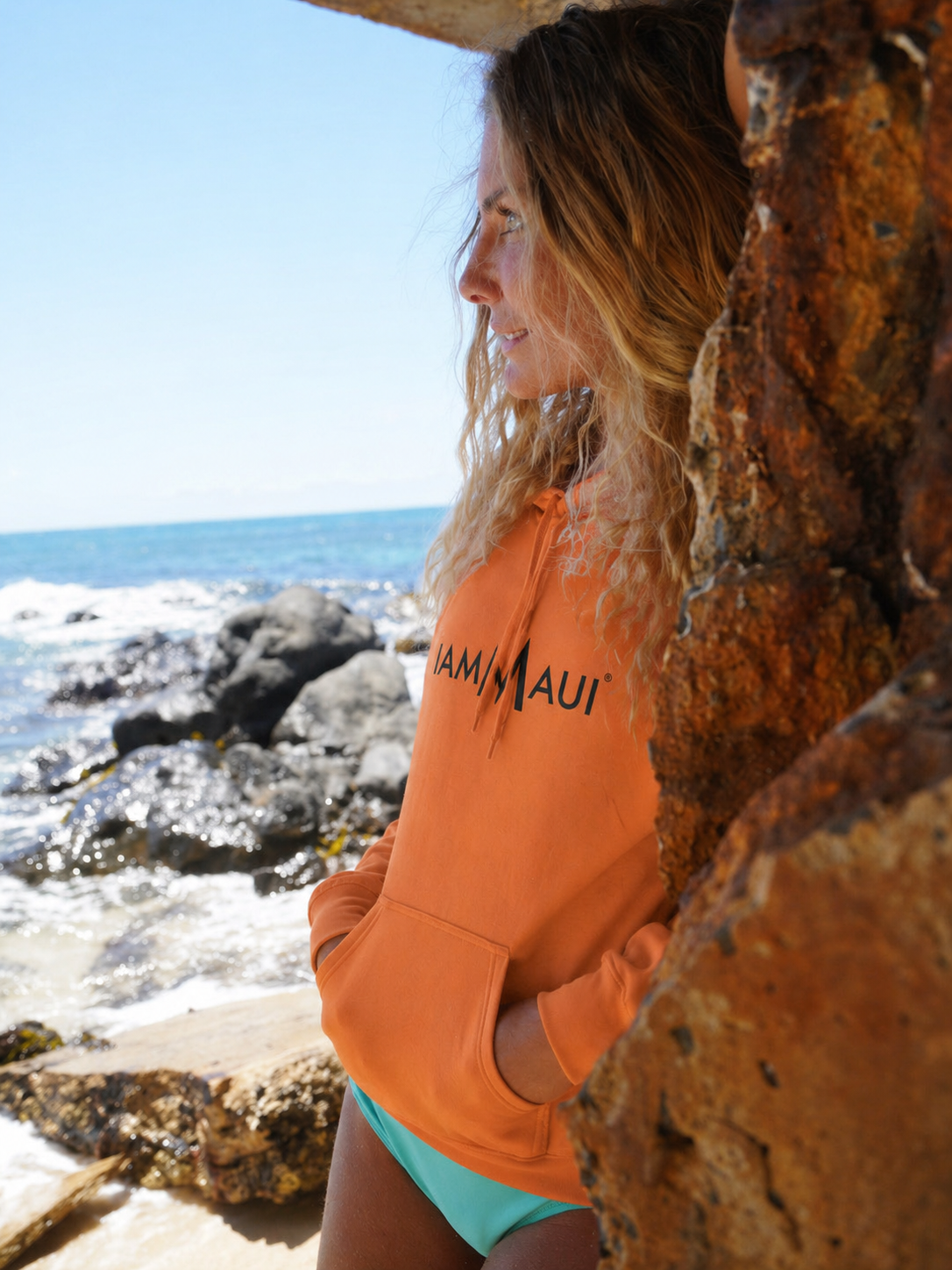 Woman wearing an orange IAMMAUI® hoodie beside the Maui ocean.